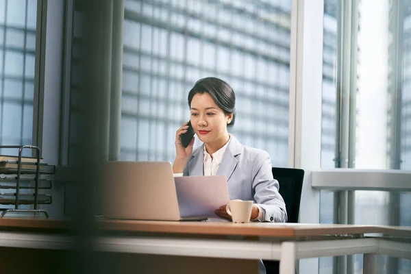 現代のオフィスでノートパソコンや携帯電話を使って仕事をしている若いアジア人のビジネスマンは — ストック写真