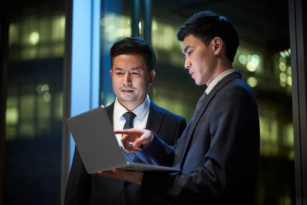 两名亚洲企业高管站在办公室窗边 讨论如何在晚上使用笔记本电脑开展业务 — 图库照片