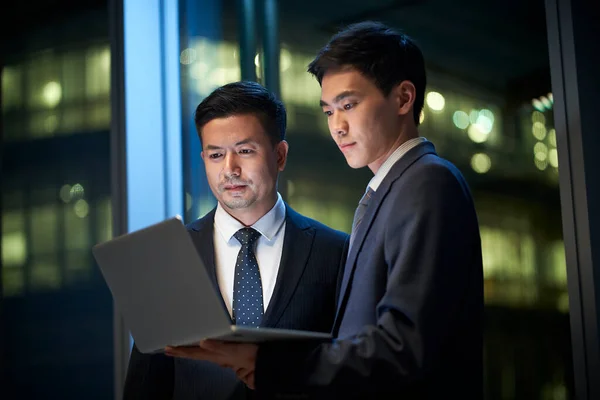 两名亚洲企业高管站在办公室窗边 讨论如何在晚上使用笔记本电脑开展业务 — 图库照片