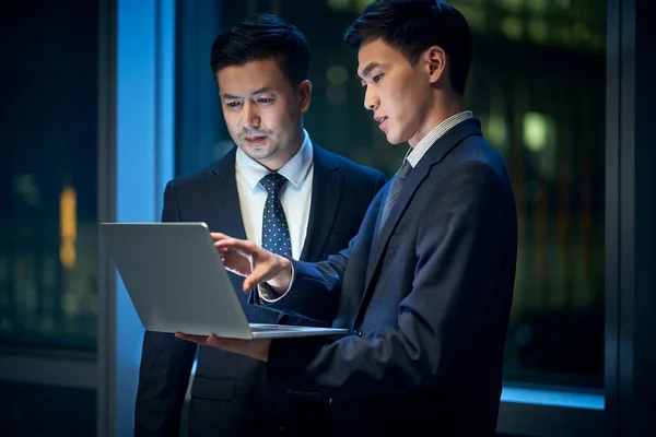 オフィスの窓のそばに立つアジア系の2人の経営者が夜にノートパソコンを使ったビジネスについて話し合い — ストック写真