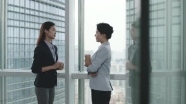 İki Asyalı iş kadını modern ofiste pencerenin önünde dikilip sohbet ediyorlar.