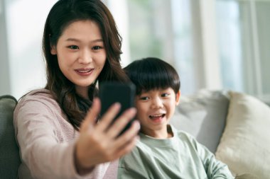 Mutlu Asyalı anne ve oğul evdeki aile koltuğunda oturup cep telefonuyla selfie çekiyorlar.