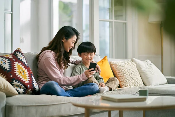 幸せなアジアの母親と息子が一緒に携帯電話の写真を見て家族のソファに座って — ストック写真