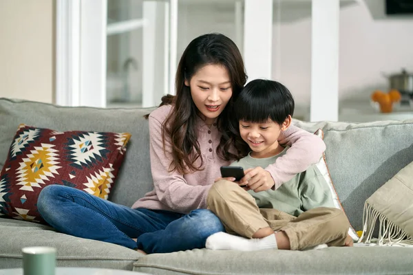 幸せなアジアの母親と息子が一緒に携帯電話の写真を見て家族のソファに座って — ストック写真