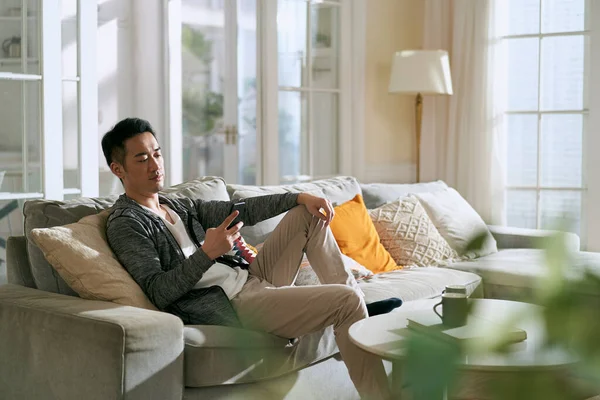 年轻的亚裔男子坐在家中客厅的沙发上 看着手机 — 图库照片