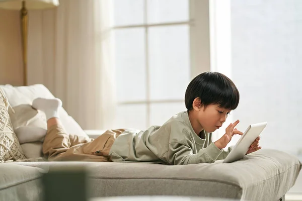 5歳の小さなアジアの少年は デジタルタブレットを使用してコンピュータゲームをプレイしている家族のソファの上に横たわっていた — ストック写真