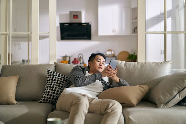 若いアジア系の成人男性は自宅で携帯電話を見てソファに横たわっ スマートフォンやソーシャルメディア中毒のための概念 — ストック写真