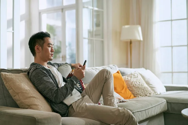 若いアジア系の成人男性が自宅のリビングでソファに座って携帯電話を見て — ストック写真