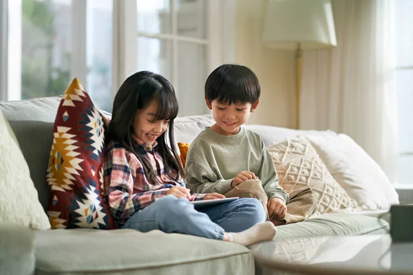 小さなアジア人の子供たち兄弟姉妹は家族のソファに座ってデジタルタブレットコンピュータを使って家にいます — ストック写真