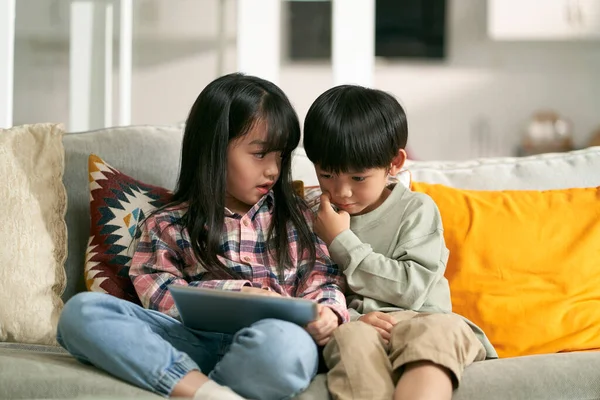 小さなアジア人の子供たち兄弟姉妹は家族のソファに座ってデジタルタブレットコンピュータを使って家にいます — ストック写真