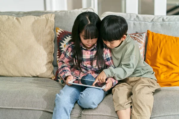 two asian children brother and sister sitting on family couch at home playing computer game using digital tablet