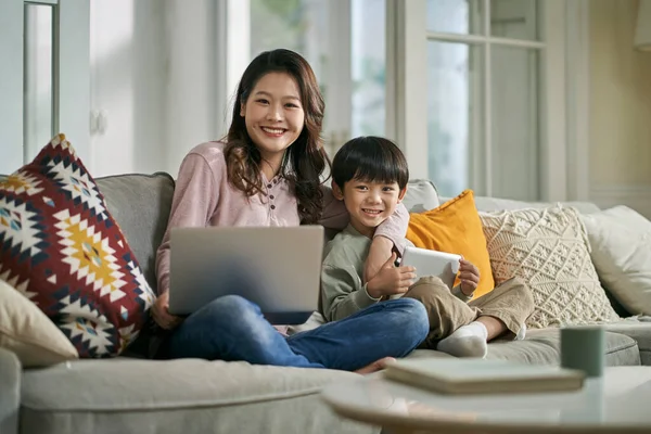 美しいアジア系の母親と5歳の息子が家でソファに座ってカメラを見て笑っていました — ストック写真