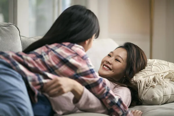 7歳の娘と楽しい会話を交わす家族のソファに寝そべっている若いアジア系の母親 — ストック写真