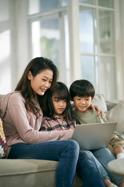 若いアジア系の母親と2人の子供が一緒にノートパソコンを使って家のソファに座って — ストック写真