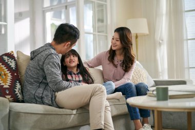 Genç Asyalı anne ve baba evdeki aile koltuğunda oturup kızlarıyla hoş sohbetler ediyorlar.