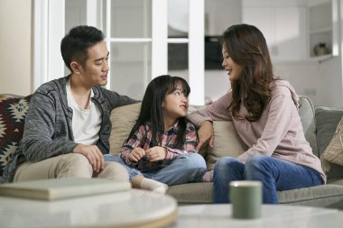 Genç Asyalı anne ve baba evdeki aile koltuğunda oturup kızlarıyla hoş sohbetler ediyorlar.