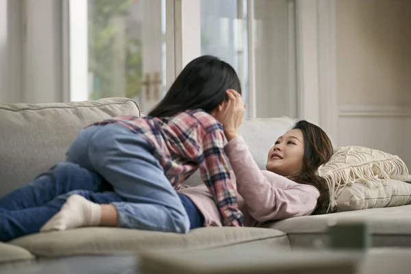 年轻的亚洲母亲躺在家庭沙发上 与7岁的女儿面对面地进行着愉快的交谈 — 图库照片