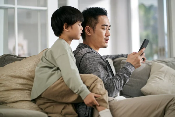 若いアジア系の父親と息子が一緒に携帯電話を見て家でソファに座って — ストック写真
