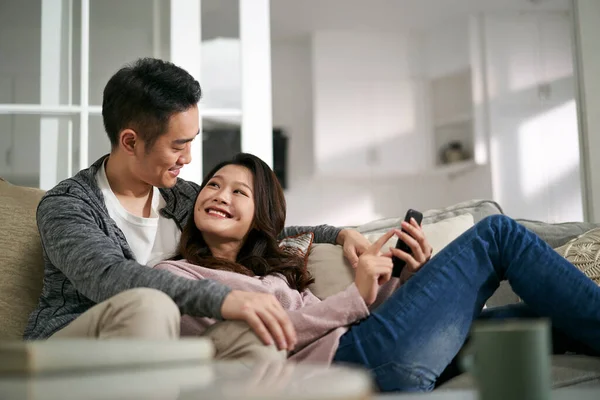 幸せな若いアジアのカップルが家でソファに座って携帯電話の写真を一緒に見て — ストック写真