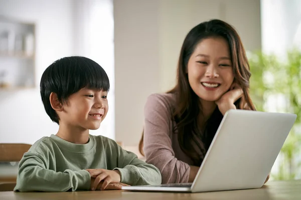 幸せなアジア系の母親と息子はノートパソコンを使って家庭用映画を見ながらテーブルに座り — ストック写真