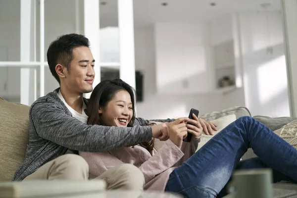 幸せな若いアジアのカップルが家でソファに座って携帯電話の写真を一緒に見て — ストック写真