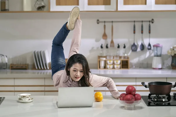 家庭のキッチンでノートパソコンを使って仕事をしながら足を伸ばす若いアジア系ビジネス女性 — ストック写真