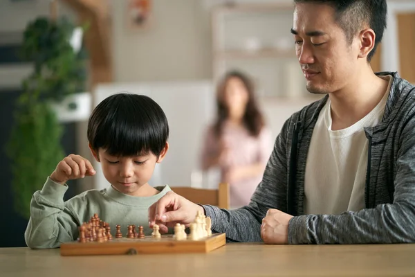 asian father and son sitting at kitchen table at home playing chess