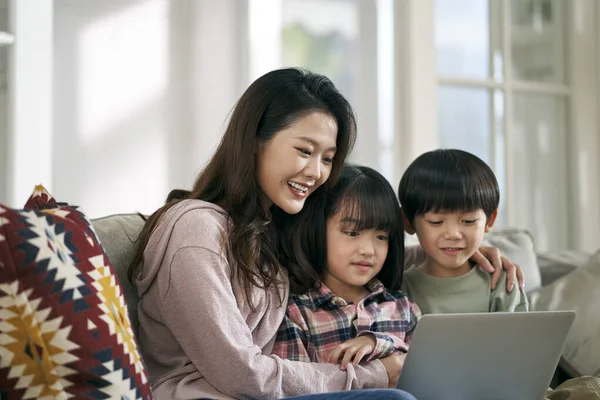 若いアジア系の母親と2人の子供が家庭用のソファに座ってノートパソコンを使って映画を見ました — ストック写真