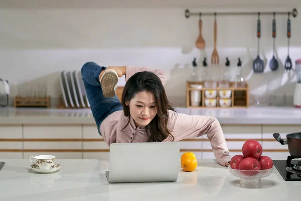 家庭のキッチンでノートパソコンを使って仕事をしながら足を伸ばす若いアジア系ビジネス女性 — ストック写真