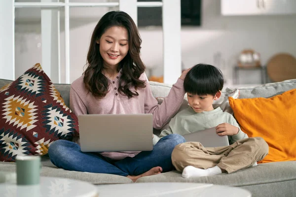 年轻的亚洲母亲和五岁的儿子坐在家里的沙发上 使用笔记本电脑和数码平板电脑 — 图库照片