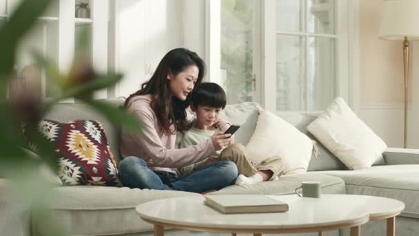 若いアジア系の母親と5歳の息子が一緒に携帯電話を見ながらソファに座って — ストック動画