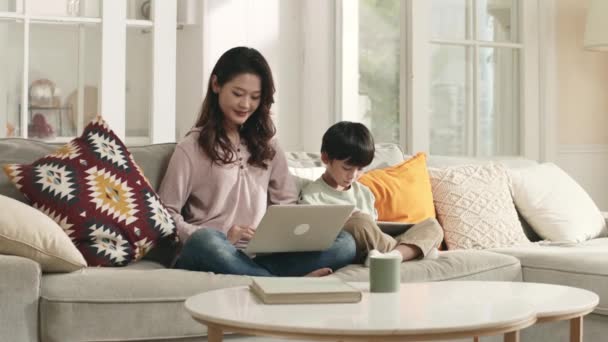 幼いアジア系の母親と5歳の息子がノートパソコンとデジタルタブレットを使って家族のソファに座り — ストック動画