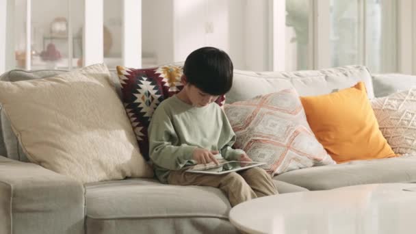 アジア系の男の子がデジタルタブレットを使ってソファでコンピュータゲームをしています — ストック動画