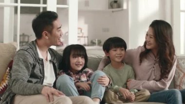 Kanepede oturup televizyon izleyen iki çocuklu mutlu Asyalı bir aile.