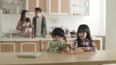 Ebeveynler aile mutfağında yemek hazırlarken iki Asyalı çocuk evde satranç oynuyor.