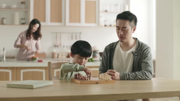 若いアジアの父親は家庭で5歳の息子とチェスをしている間 母親は家族のキッチンで食事を準備しています — ストック動画