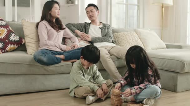 Két Ázsiai Gyerek Padlón Játszik Miközben Szülők Beszélgetnek Kanapén Háttérben — Stock videók