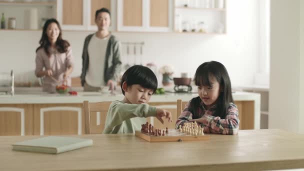 2人のアジアの子供が家のダイニングテーブルに座ってチェスをしている間 両親はバックグラウンドでキッチンで食事を準備しています — ストック動画