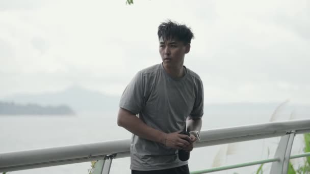 年轻的亚洲男子运动员 手里拿着水瓶 回头望着大海 — 图库视频影像