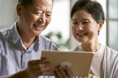 Mutlu Asyalı çift evdeki koltukta oturup dijital tablet kullanıyorlar.