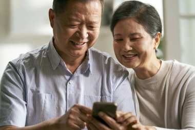 Mutlu Asyalı çift evdeki koltukta birlikte cep telefonu kullanıyor.