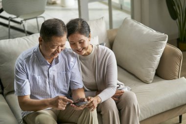 Mutlu Asyalı çift evdeki koltukta birlikte cep telefonu kullanıyor.