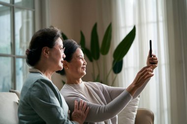 İki kıdemli Asyalı kadın evde koltukta oturup selfie çekerken mutlu bir şekilde cep telefonu kullanıyor ve gülümsüyor.