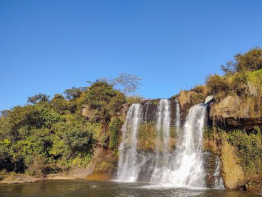 Berrak bir mavi gökyüzünün altındaki gür bitki örtüsüyle çevrili kayalık uçurumlarda çağlayan sakin bir şelale huzurlu ve huzurlu bir doğal ortamı yansıtıyor..