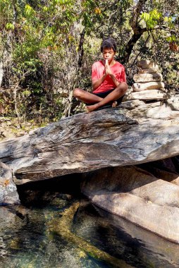 Orman deresinin yakınındaki büyük bir kayanın üzerinde meditasyon yapan genç bir çocuğun sakin bir görüntüsü. Yeşillik ve güneş ışığıyla çevrili. Doğada huzur ve farkındalık yayıyor..