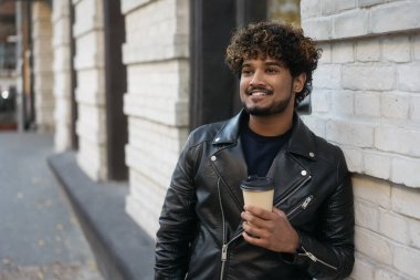 Siyah deri ceketli yakışıklı gülümseyen Hintli adam elinde bir fincan kahveyle caddeye bakıyor. Duvarın yanında duran havalı Asyalı hipster portresi sıcak içecekle, boşluğu kopyala 