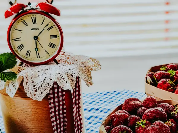 Kırmızı kartonlu masa örtüsü ve klasik çalar saati olan karton kutularda taze çileklerin dikey kırsal sahnesi.