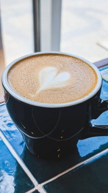 Kalp şeklindeki latte sanatıyla kaplı sıcak kapuçino dolu siyah bir seramik fincan.