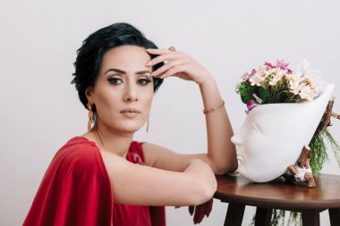 Kırmızı elbiseli düşünceli genç bir kadın, düşünceli ve stüdyoda çiçekli alçıdan bir vazoyla poz veriyor.