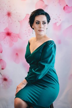 Pembe çiçekli arka planda, sandalyede oturan lüks bir kadının dikey bahar tarzı stüdyo portresi.